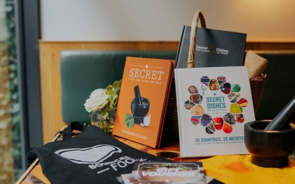 three bounceback food cookbooks propped up on a table, with a tea towel and a apron