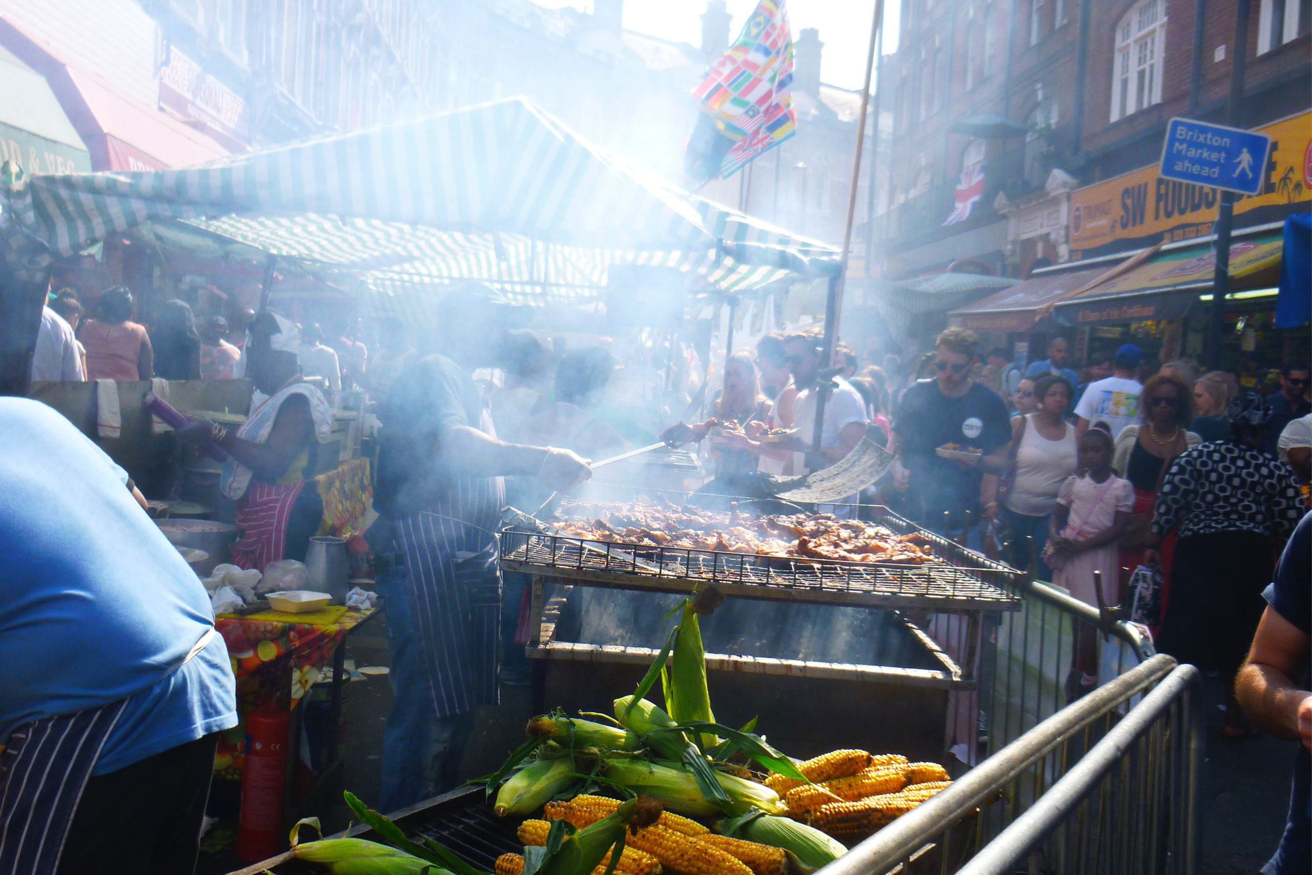 Brixton food market. There is a barbecue with smoke rising from it and fresh corn. The market is crowded with people browsing.