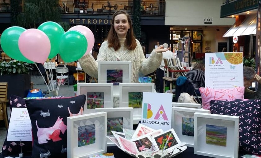 A volunteer for Bazooka Arts stood at a market stall with balloons in the background.