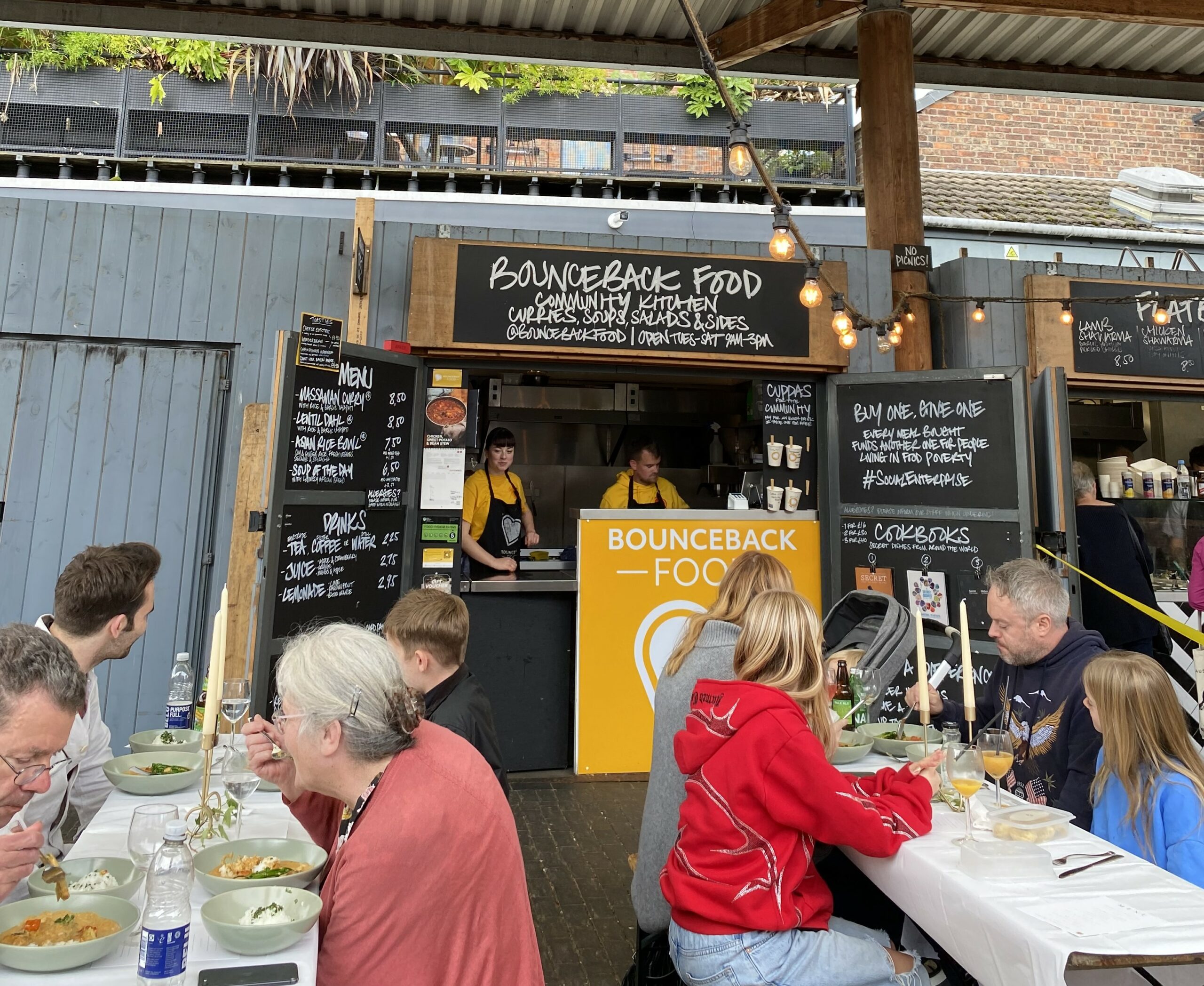 People are eating at tables in front of the Bounceback Food Community Kitchen.